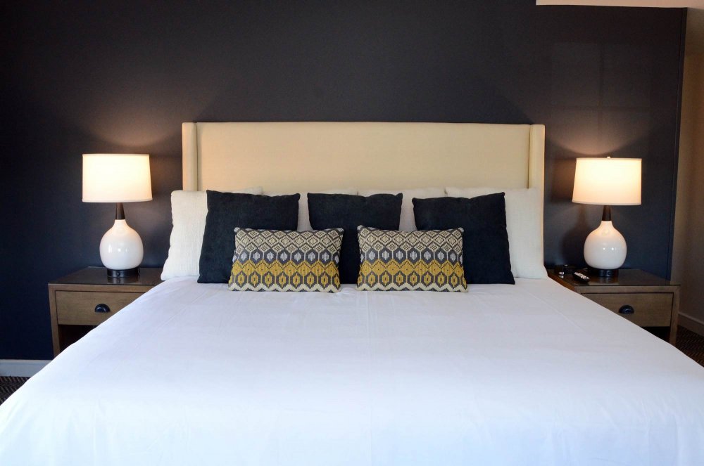 A made bed at a historic boutique hotel in Elberton, GA, with a tall cream headboard, dark accent pillows, and patterned throw pillows, flanked by two nightstands with matching lamps.