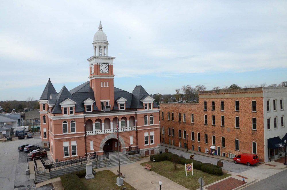 An aerial view of a three-story peach-colored courthouse with a clock tower, situated in an Elberton, GA town square next to a multi-story brick historic boutique hotel.