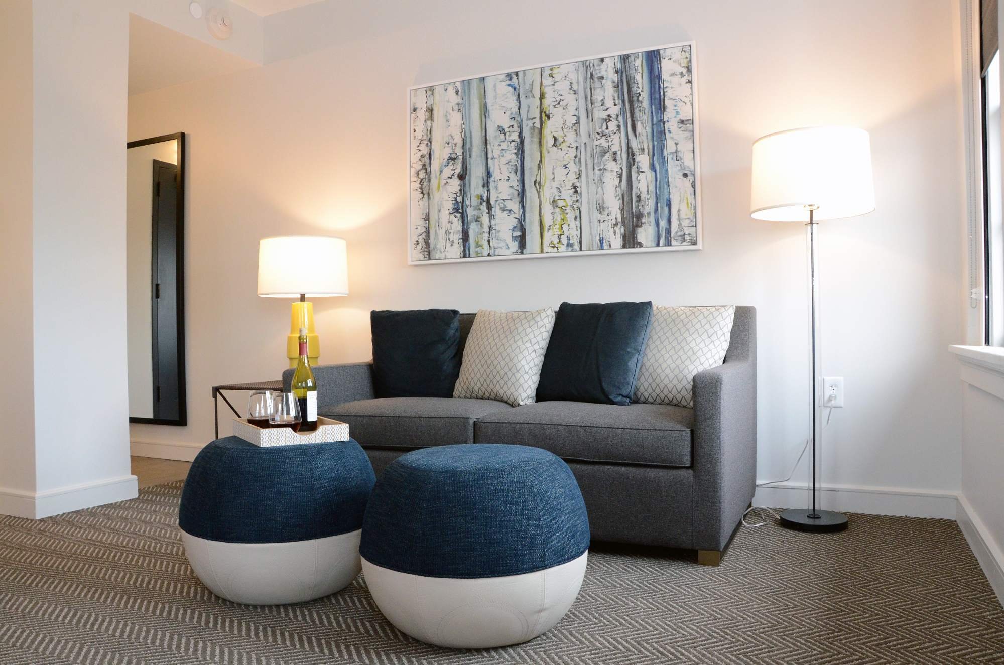 A gray sofa with navy and patterned pillows, two circular blue and white ottomans, and a wine tray in a living room at a historic boutique hotel in Elberton, GA.