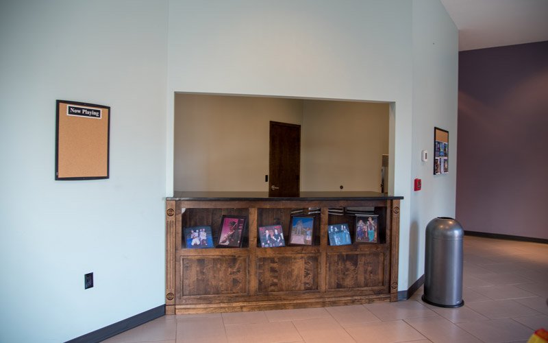 A service counter in the Elberton Arts Center lobby with framed photographs along the front, a “Now Playing” corkboard on the left wall, and a trash receptacle on the right.