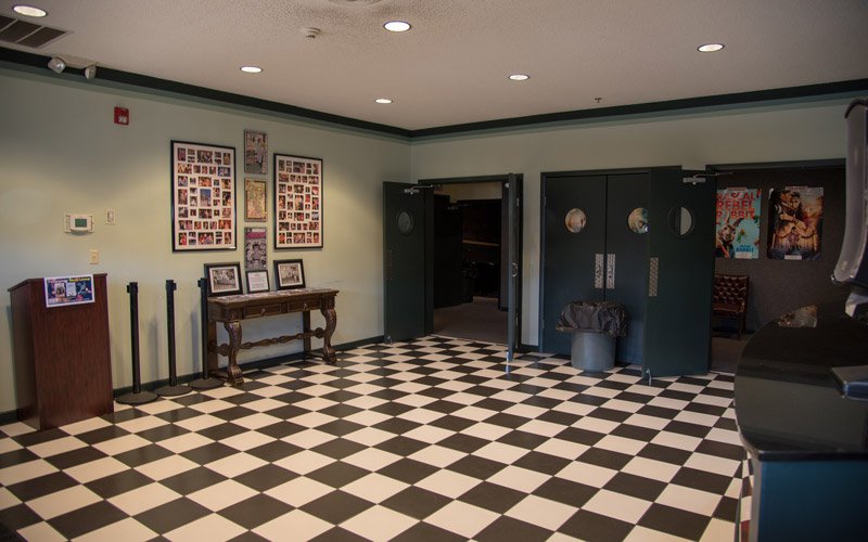 The lobby of the Elbert Theatre in Elberton, GA, featuring a black and white checkered floor, a wooden console table with framed photos, and two dark green double doors leading into the auditorium.