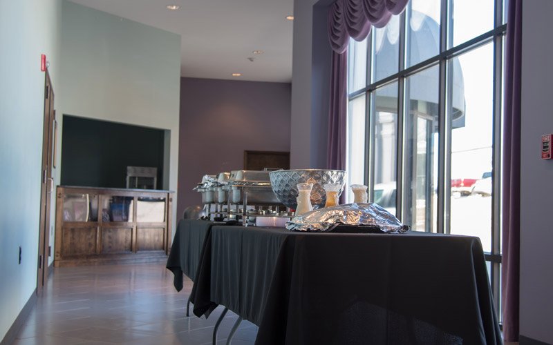 A black-clothed buffet table with chafing dishes and serving bowls set up in front of a large window at the Elberton Arts Center in Elberton, GA.