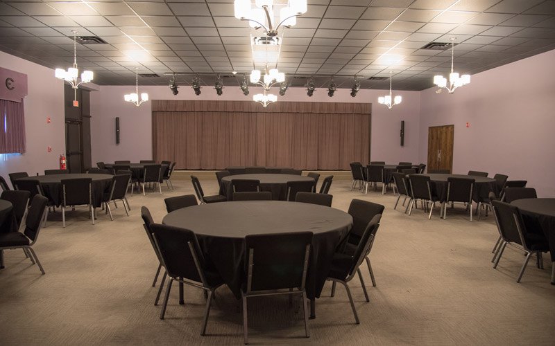 A banquet hall at The Elberton Arts Center featuring multiple round tables with black tablecloths, surrounded by chairs, facing a stage with a drawn curtain.